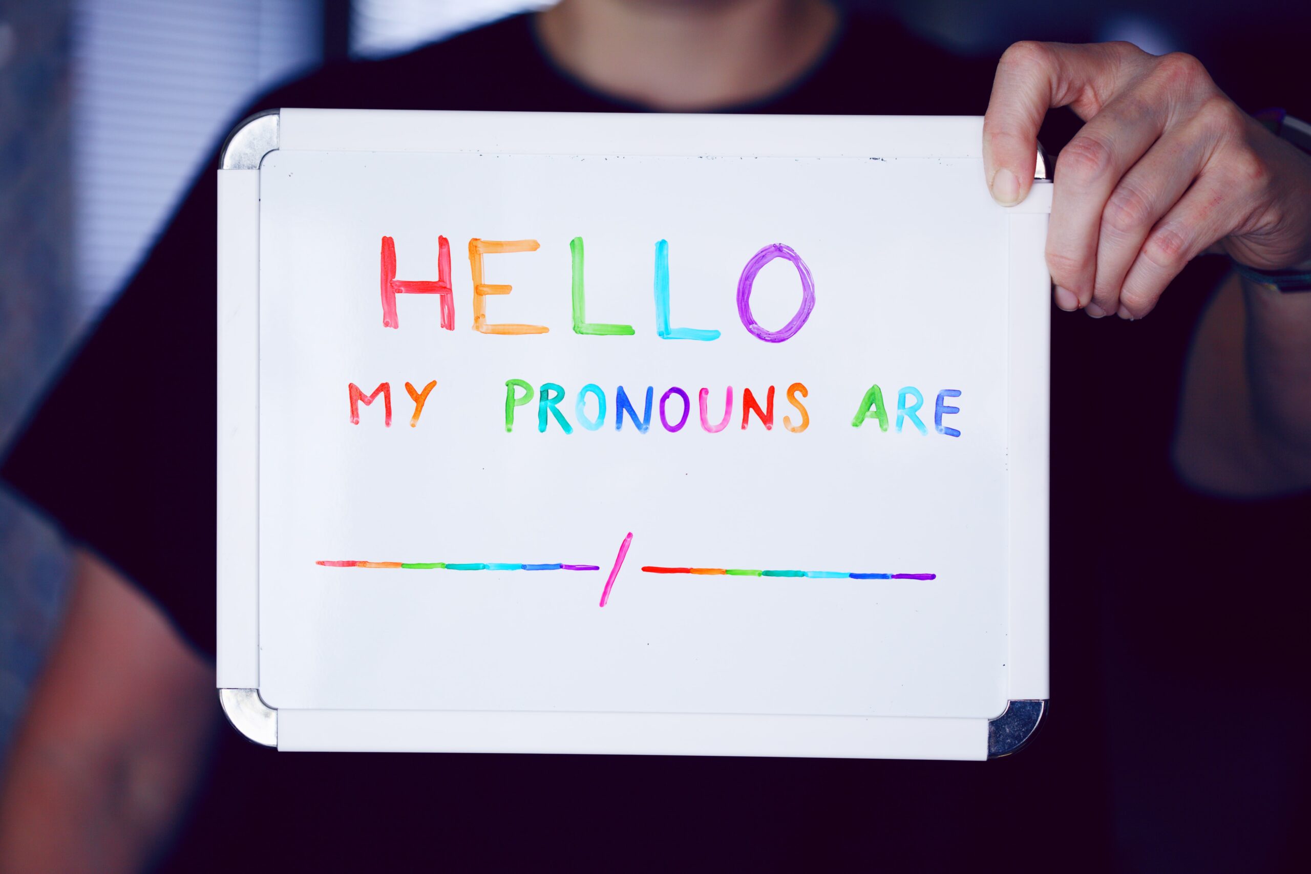 Hello. My pronouns are ___/___ (written in rainbow colors on dry erase board that a faceless person is holding)